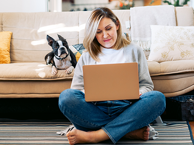 woman sitting on floor doing remote work on laptop with dog on couch behind her