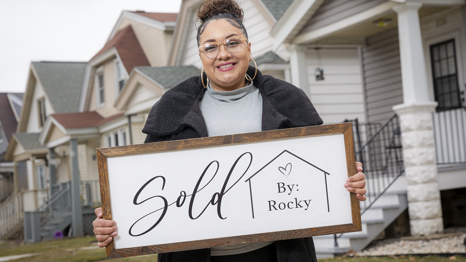 Raquel Aleman holds up a Sold by Rocky sign in front of houses